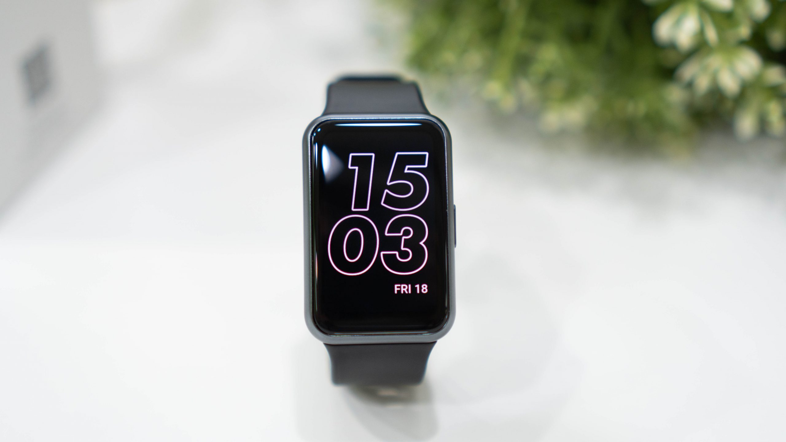  A black Huawei Watch Fit 3 with a black silicone strap is sitting on a solid white surface with a green plant out of focus in the background.